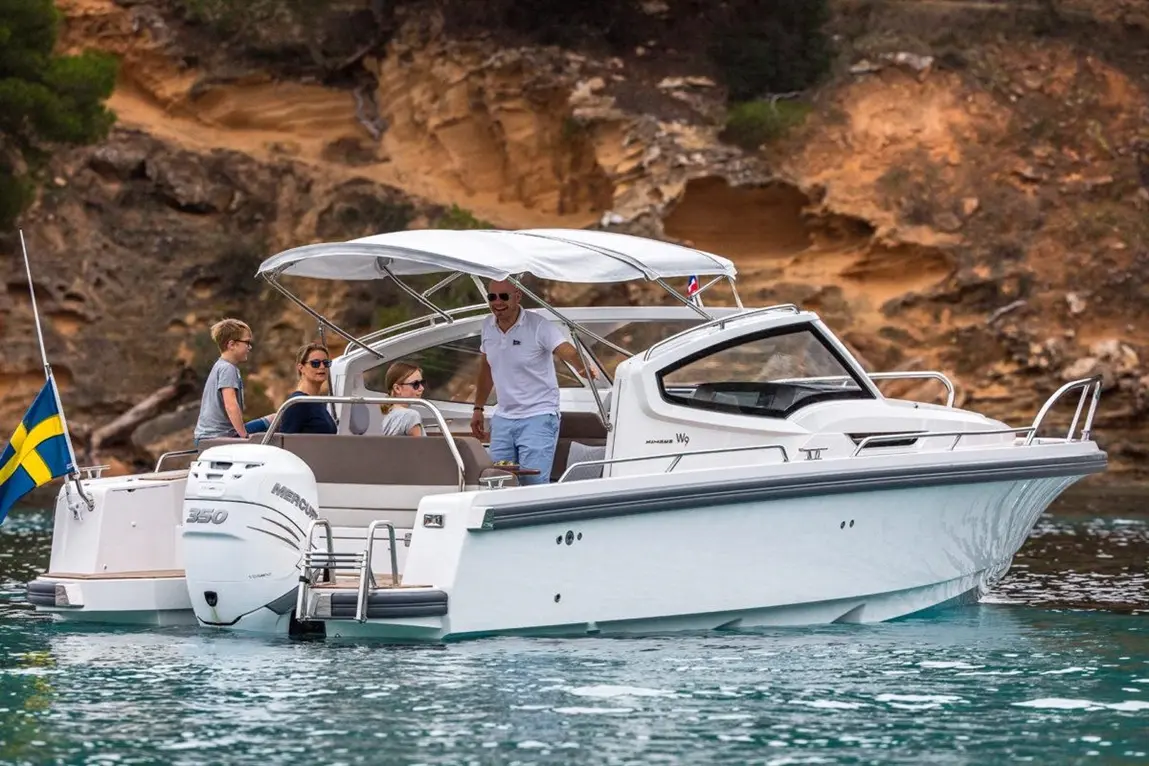 Family enjoying a boat ride on a white motorboat with a Swedish flag, green hills and rocky shoreline in the background.